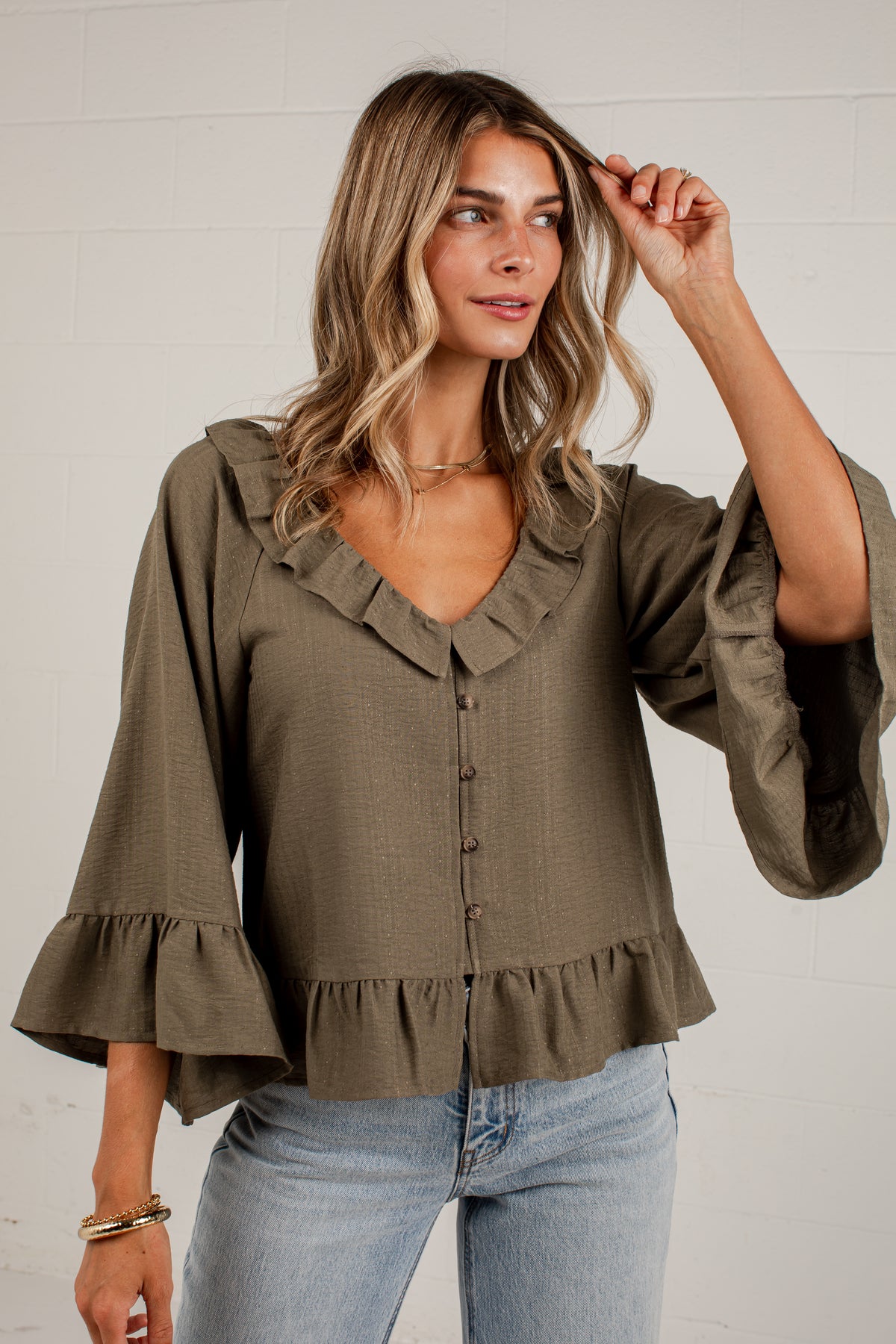 A woman wearing an olive green blouse with ruffled trim, button-front detail, and flared bell sleeves, paired with light blue jeans, standing against a white brick wall.