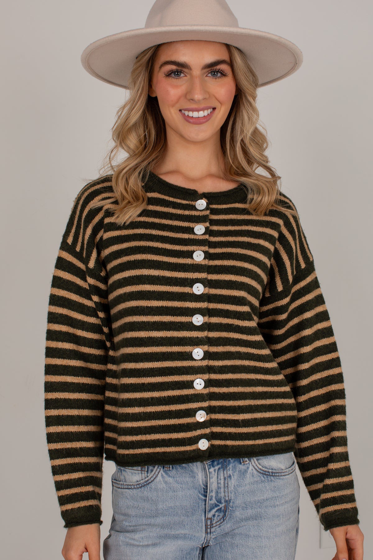 A woman wearing a cropped, long-sleeve knit cardigan with horizontal black and tan stripes, featuring a button-down front. She is also wearing a wide-brimmed beige hat and light-wash blue jeans, photographed against a solid light gray studio background.