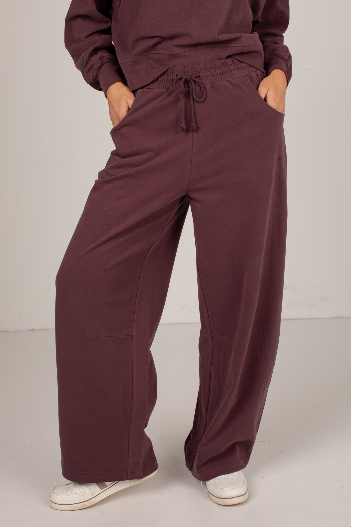 A person wearing loose, wide-leg burgundy pants with a drawstring waistband and side pockets, paired with white sneakers, shown from the waist down against a plain light gray studio background.