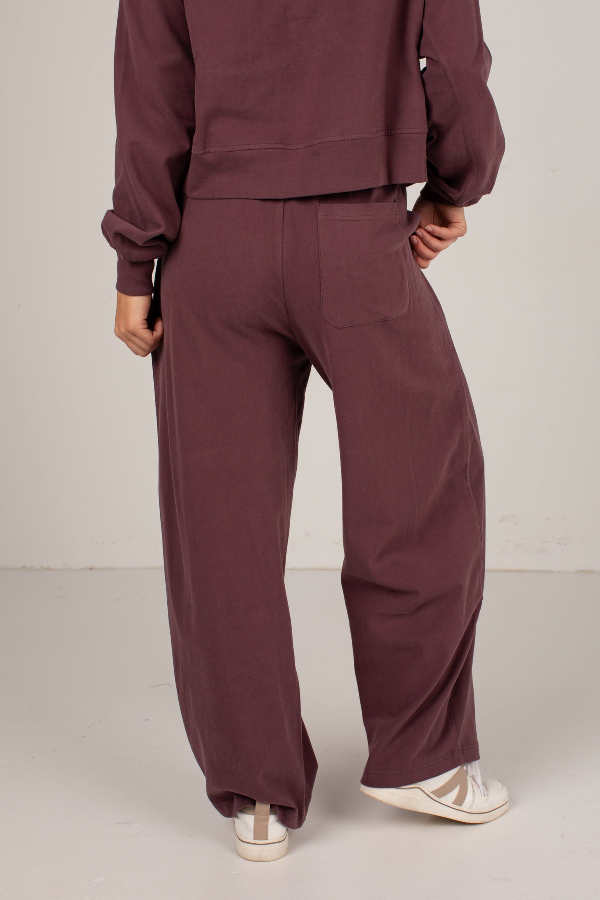 A person shown from the back wearing a matching mauve-brown sweatshirt and wide-leg sweatpants, paired with white sneakers, against a plain light gray studio background.