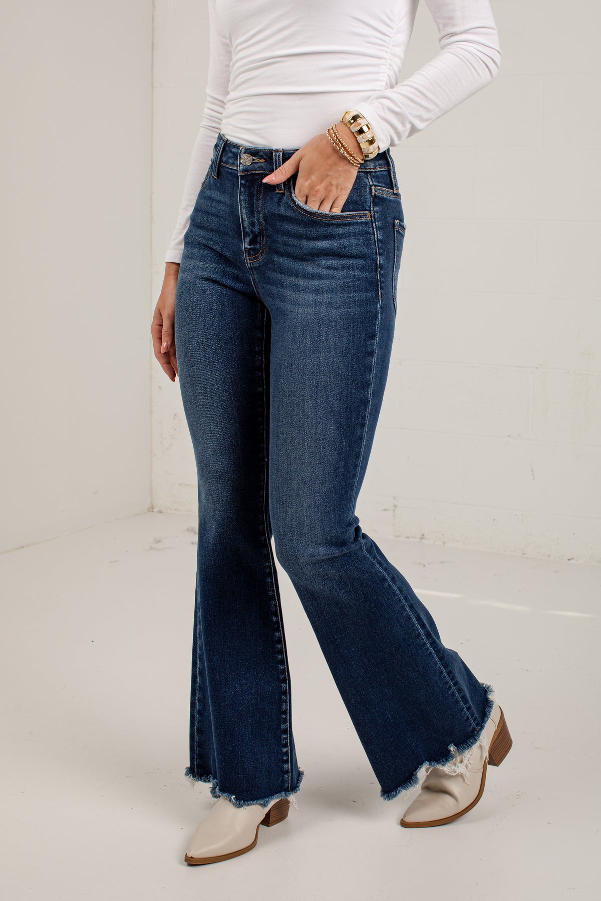 A model wearing dark blue high-waisted flare jeans with a frayed hem, paired with a white long-sleeve top and white western-style boots, standing against a plain off-white wall.