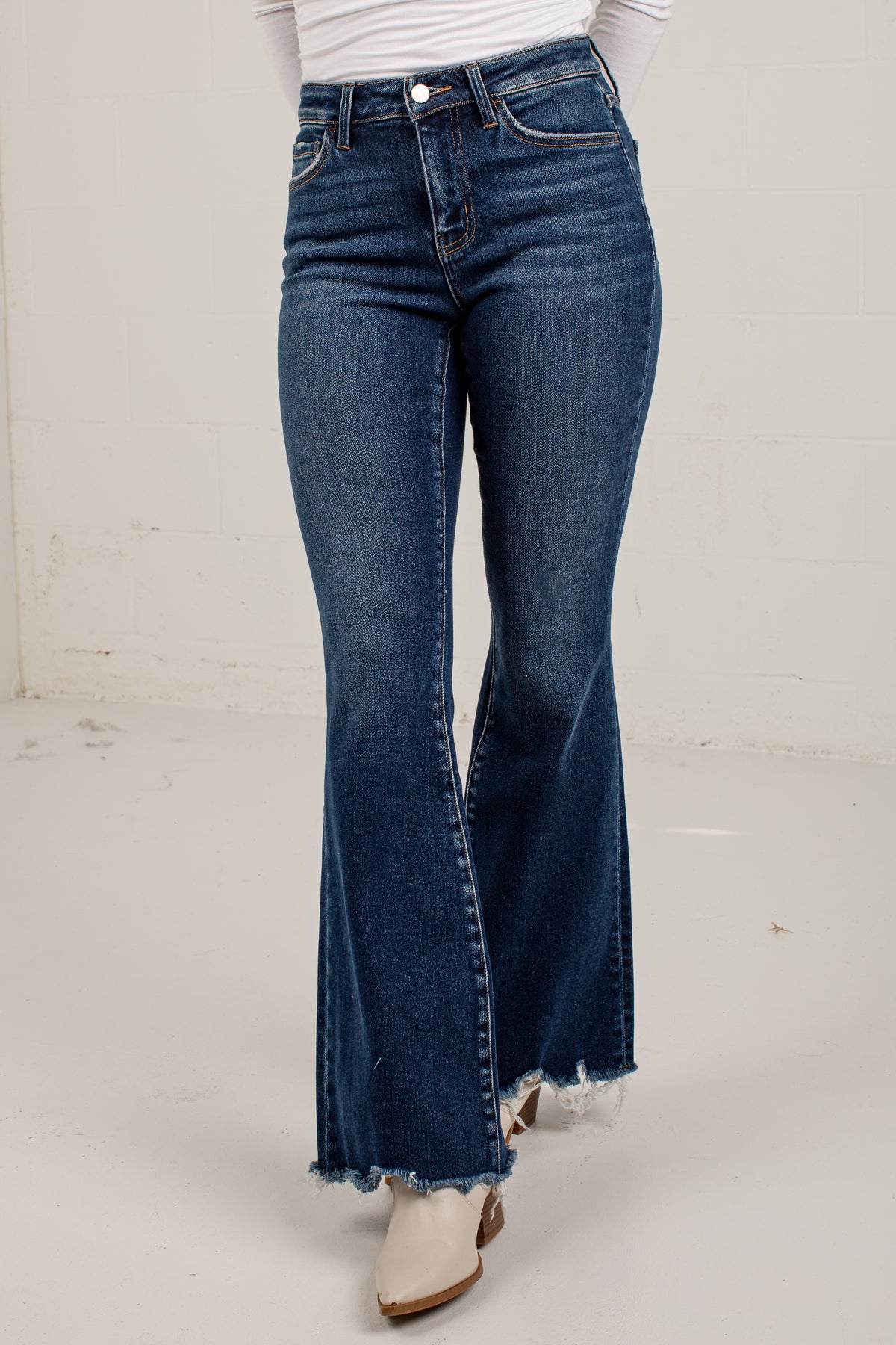 A person wearing dark blue flare jeans with a frayed hem, paired with a white top and white boots, standing against a plain off-white wall.