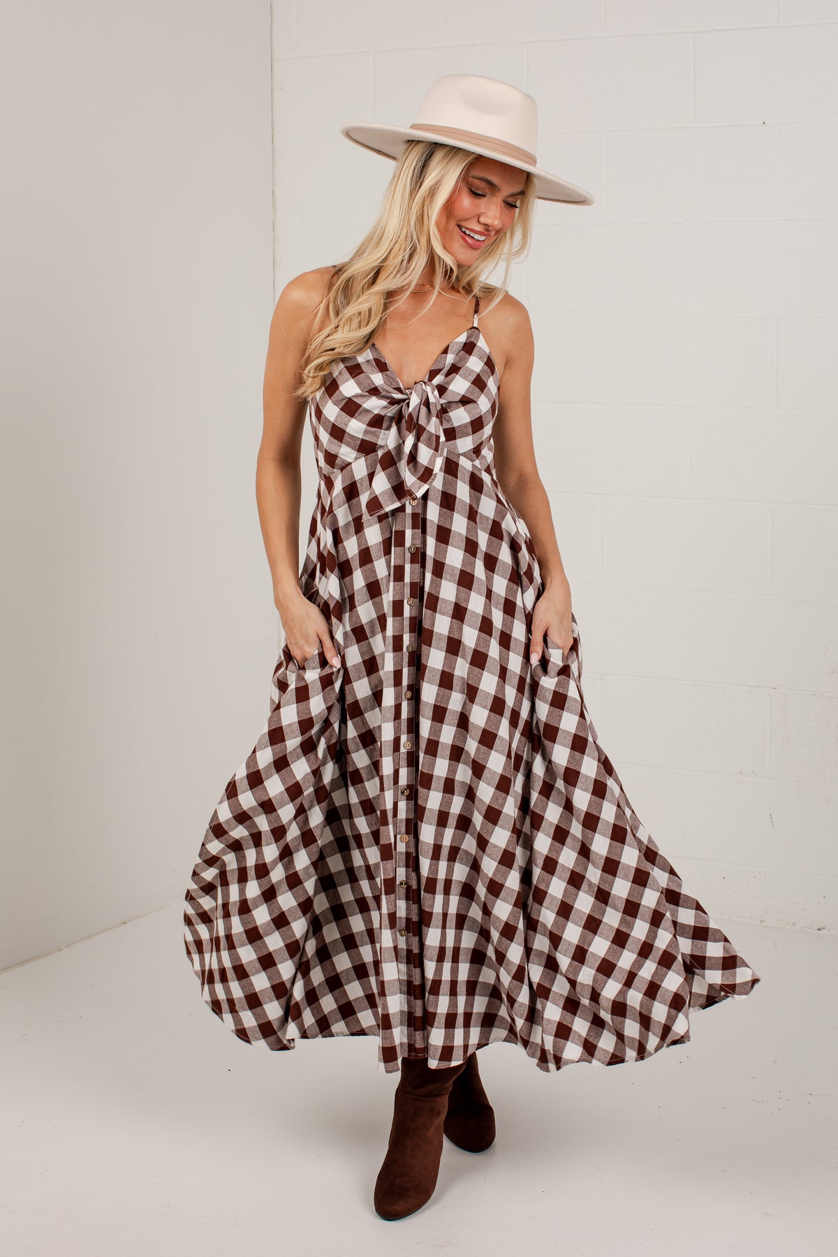 A woman modeling a brown and white gingham maxi dress with a tie-front bodice and pockets, styled with a cream wide-brim hat and brown ankle boots against a white studio background.