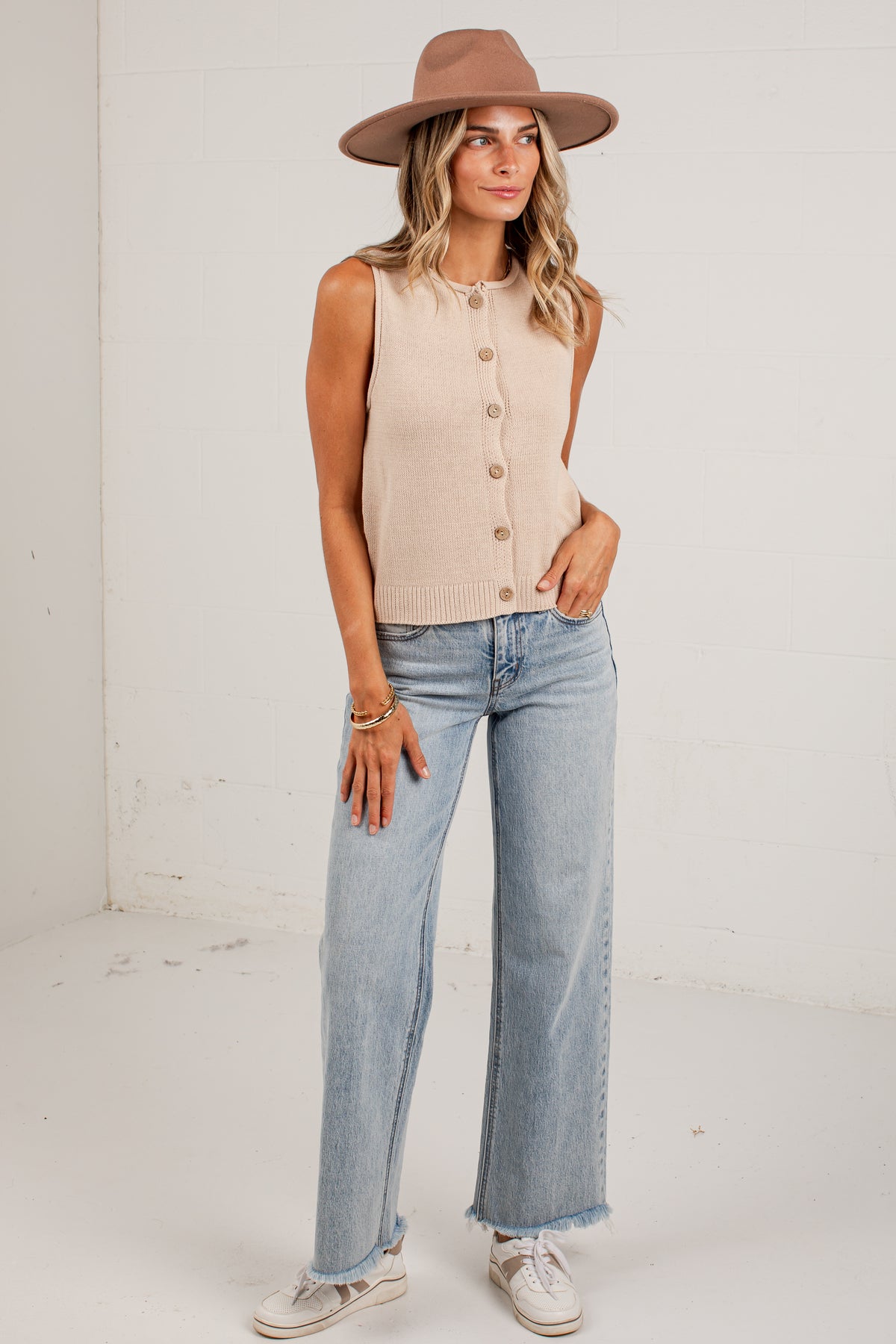 A woman modeling a sleeveless beige knit button-front top with light-wash wide-leg jeans, a brown wide-brim hat, and white sneakers, standing against a plain white wall.