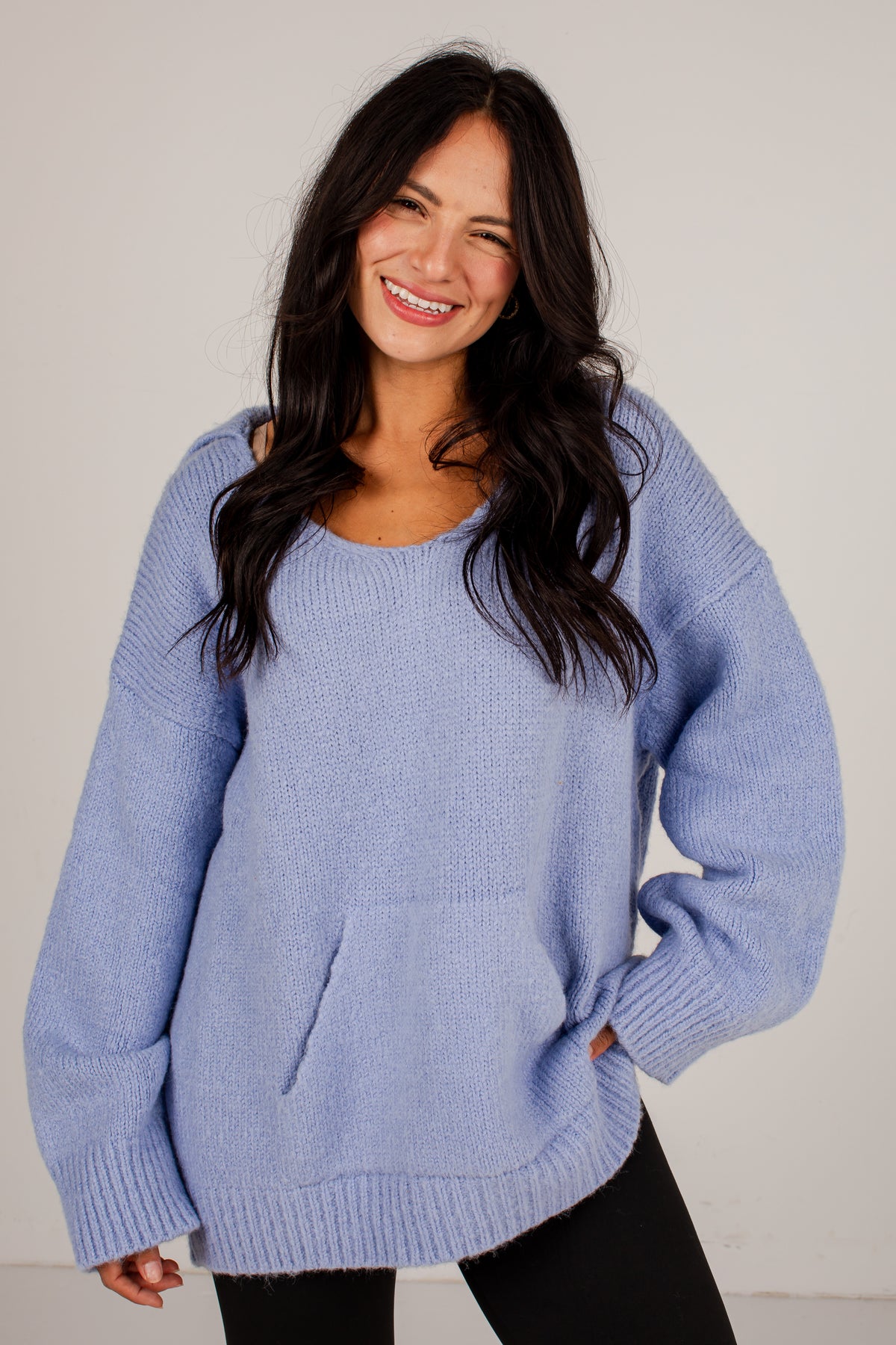 A woman wearing a light periwinkle blue knit sweater with a crew neck and front pockets, paired with black pants, against a plain light gray background.