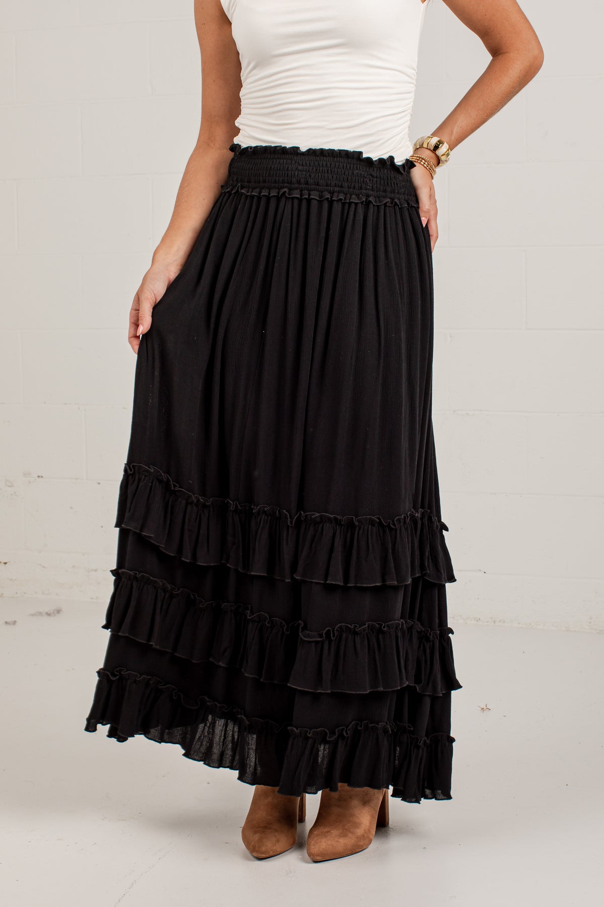 A black tiered maxi skirt with an elastic waistband, worn by a model with a white top and brown suede boots against a white brick wall.