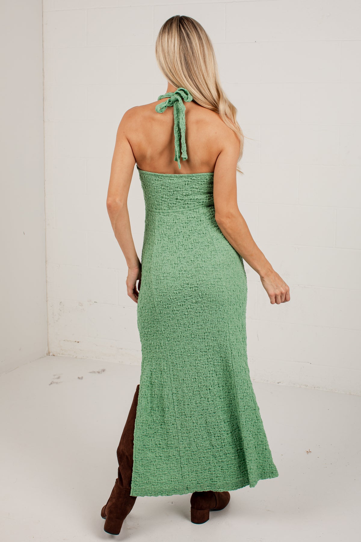 A woman shown from the back wearing a textured sage green halter-neck maxi dress with a tie at the back of the neck, paired with brown boots, in a white studio setting.