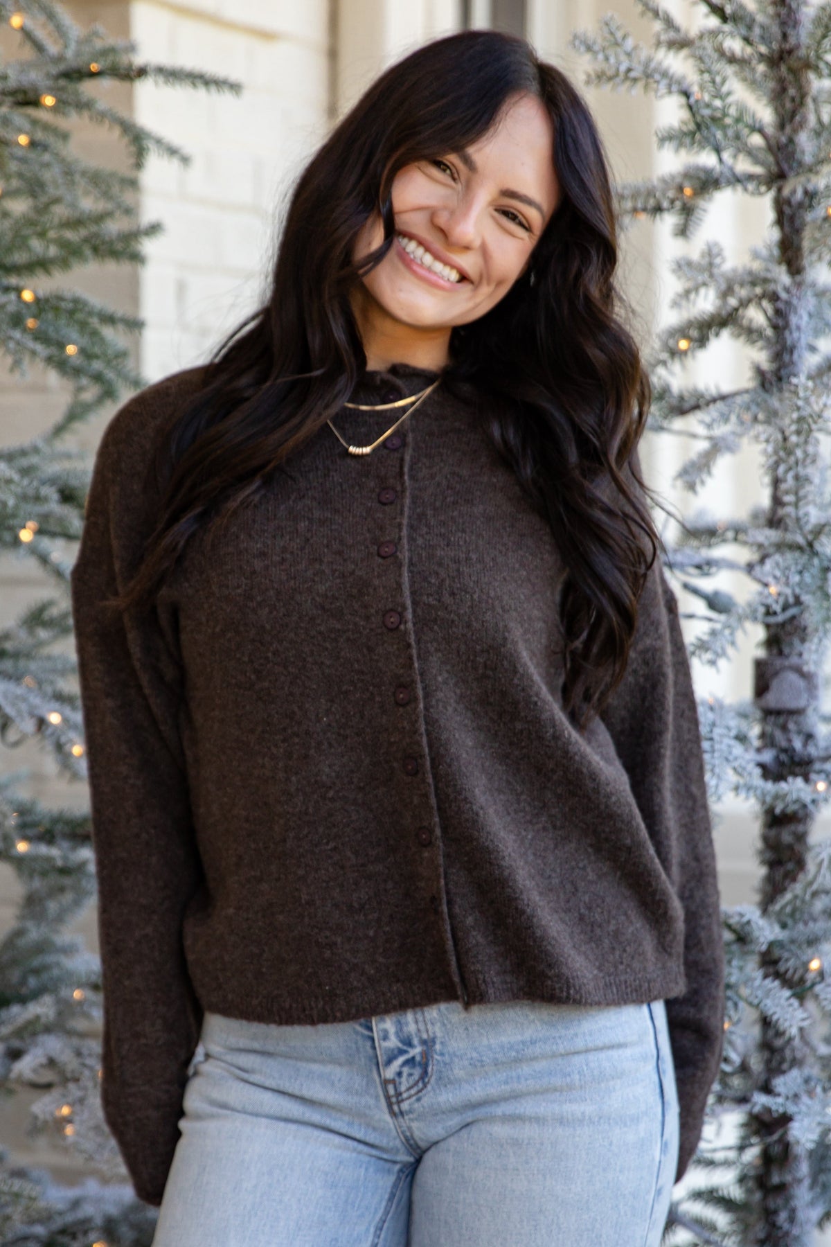 A woman wearing a dark brown knit button-front sweater and light-wash blue jeans, standing outdoors in front of decorated Christmas trees.