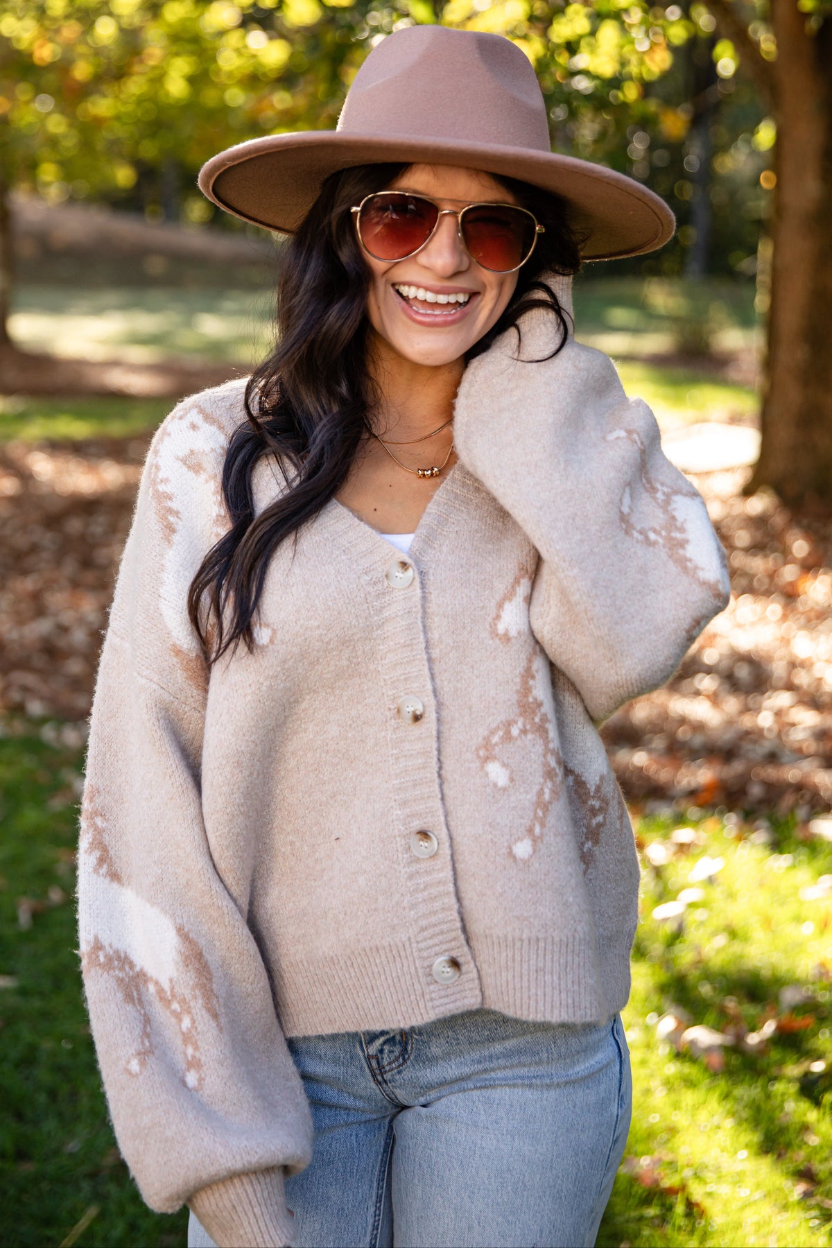 A woman outdoors wearing a beige knit cardigan with white distressed patches, a dusty pink wide-brim hat, and sunglasses, standing in a sunny park with trees and fallen leaves.