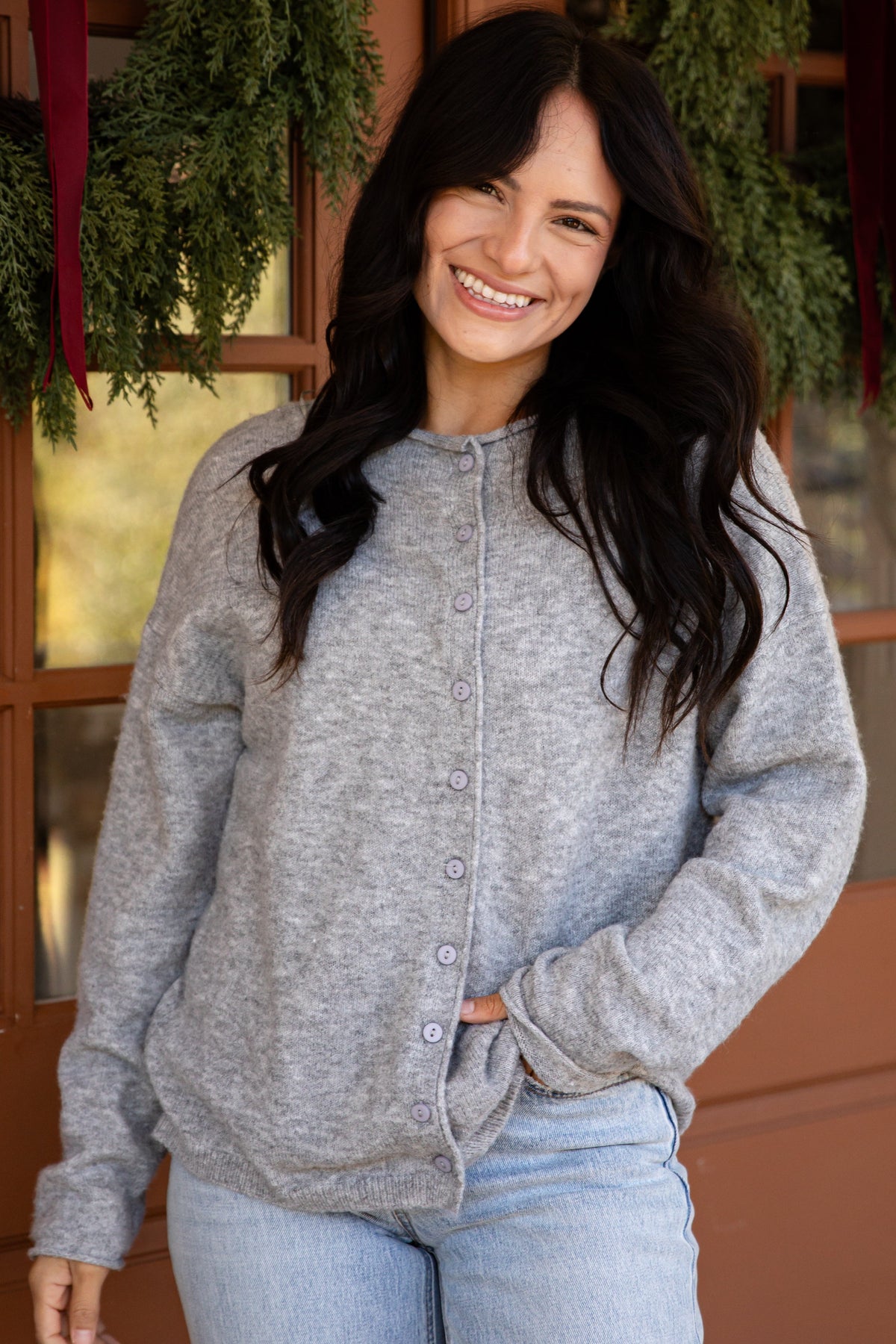 A woman smiling and wearing a light gray heathered button-front cardigan sweater and light blue jeans, standing in front of a warm brown door decorated with green garlands.