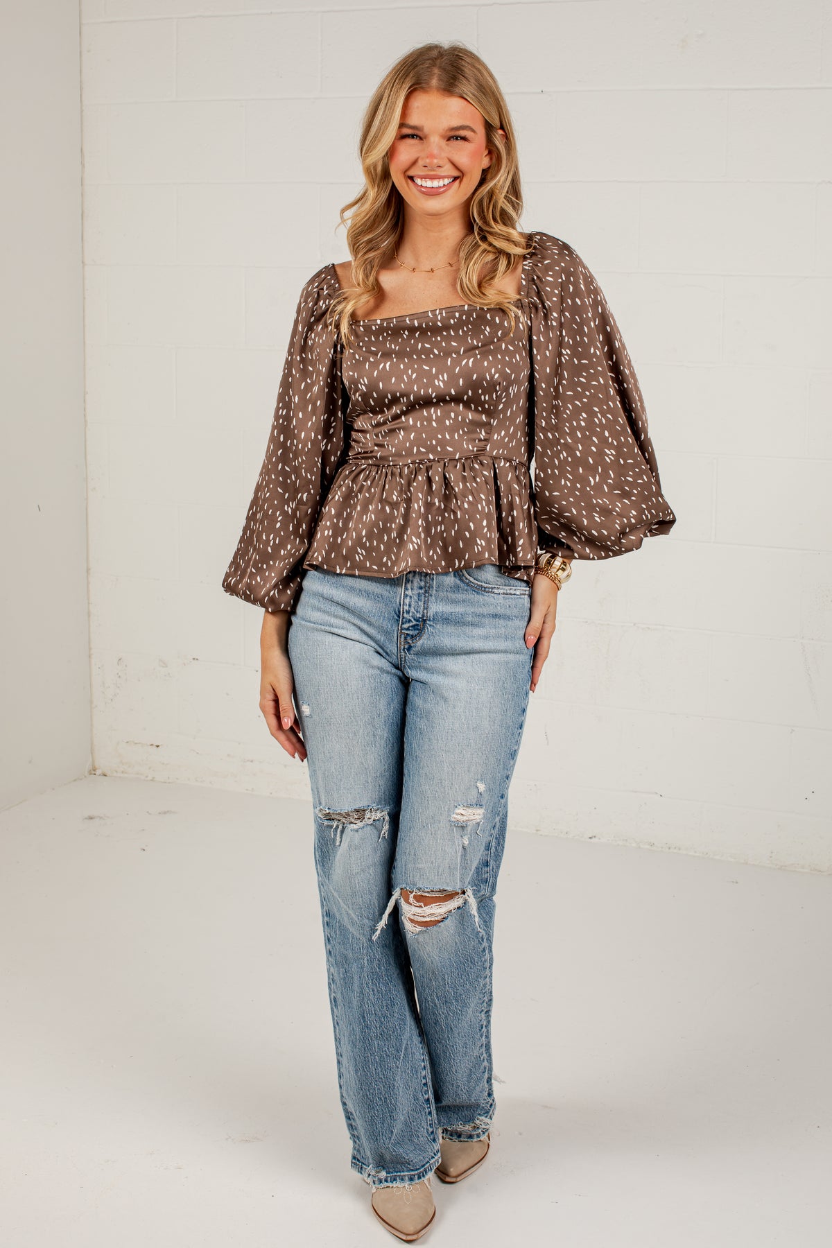 A woman modeling a brown and white patterned puff-sleeve peplum top with distressed light-wash blue jeans, standing against a white studio wall.