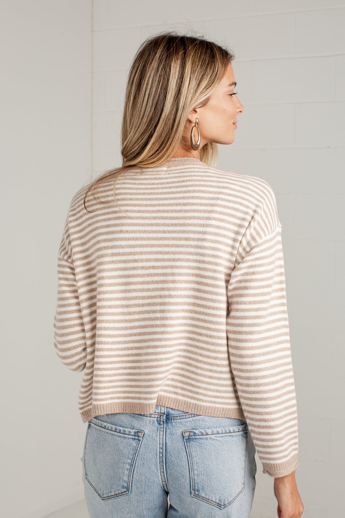 A woman shown from the back wearing a beige and white horizontally striped knit sweater and light blue jeans, standing against a white brick wall.