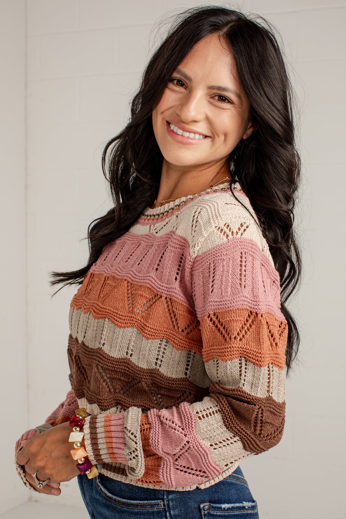 A smiling woman with long dark hair wearing a multi-colored knit sweater with horizontal stripes in pink, orange, brown, and cream, paired with blue jeans.