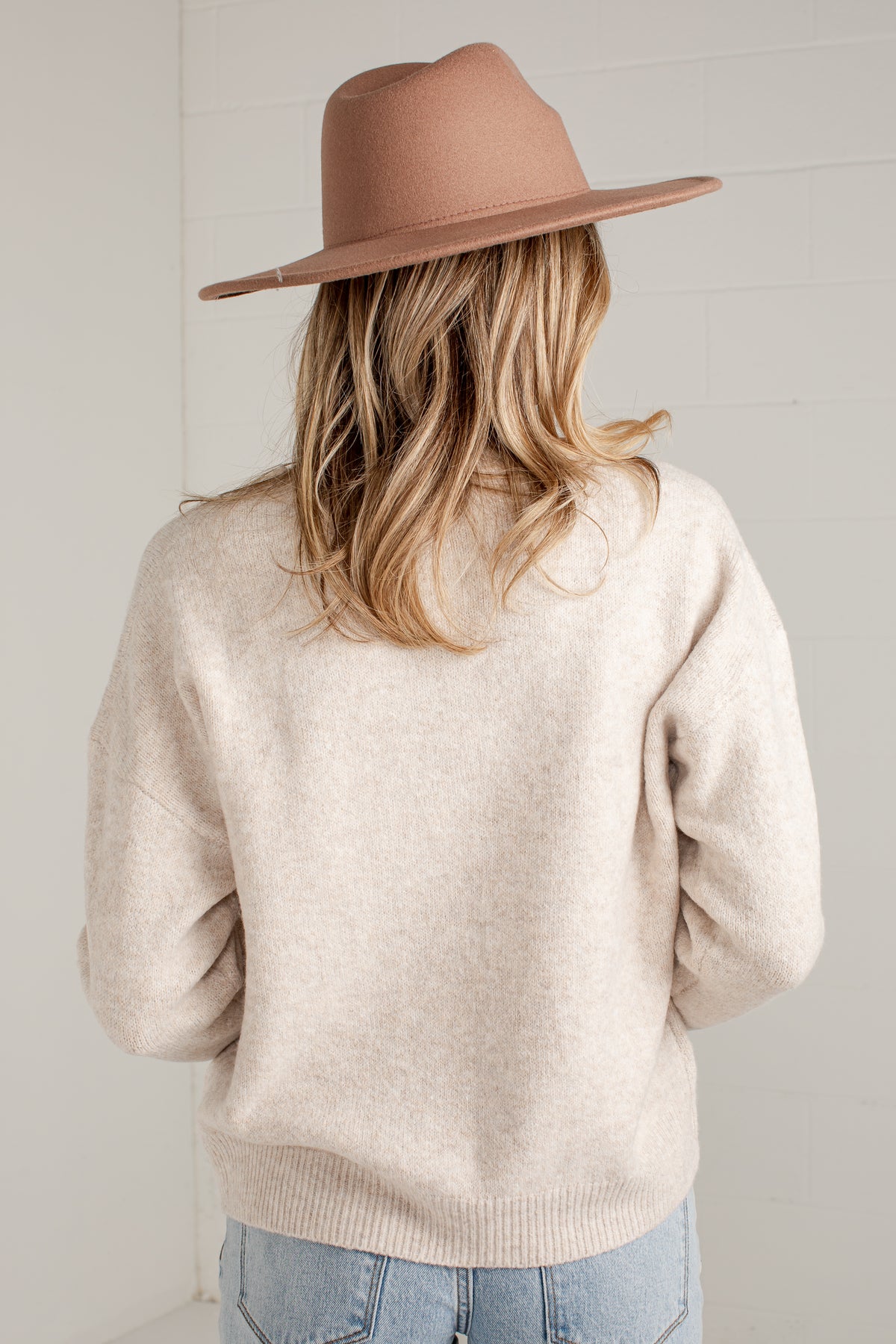 A woman shown from behind wearing a light beige knit sweater, a dusty pink wide-brim fedora hat, and light blue jeans against a white paneled wall.
