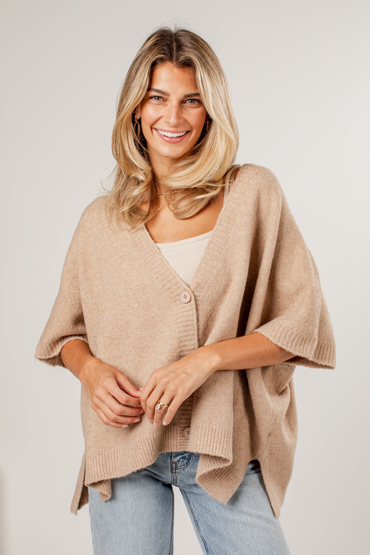 A smiling blonde woman wearing a beige knit poncho cardigan over a white top and light blue jeans, photographed against a plain light gray studio background.