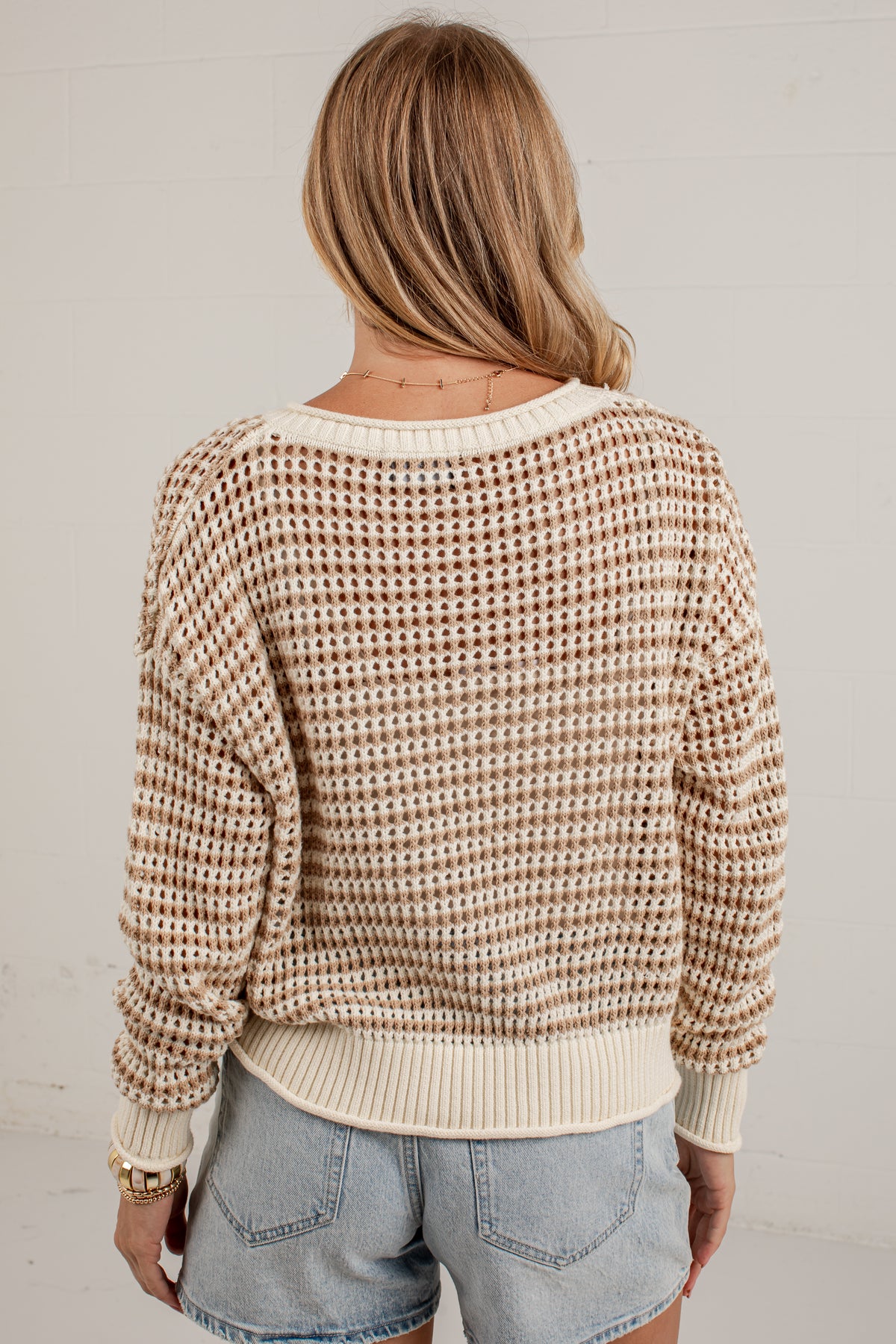 A woman viewed from behind wearing a cream and brown waffle-knit sweater with light blue denim shorts, standing against a white wall.