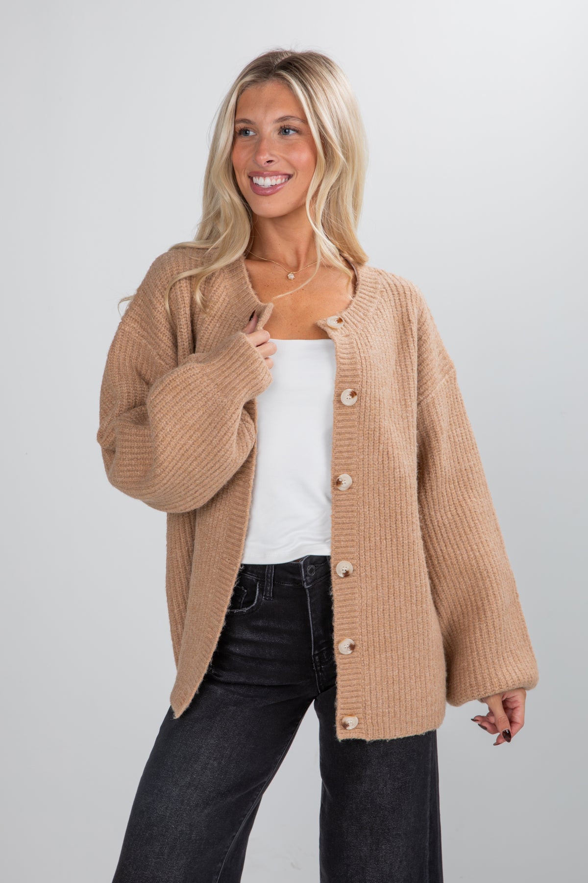 Woman wearing a beige cardigan over a white top and black pants on a plain background