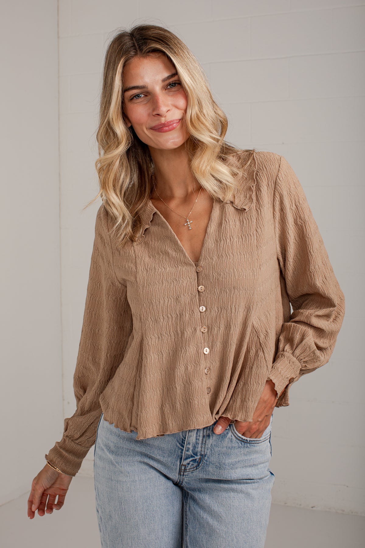 A woman wearing a light brown crinkled long-sleeve button-up blouse with light blue jeans, standing against a white paneled wall.