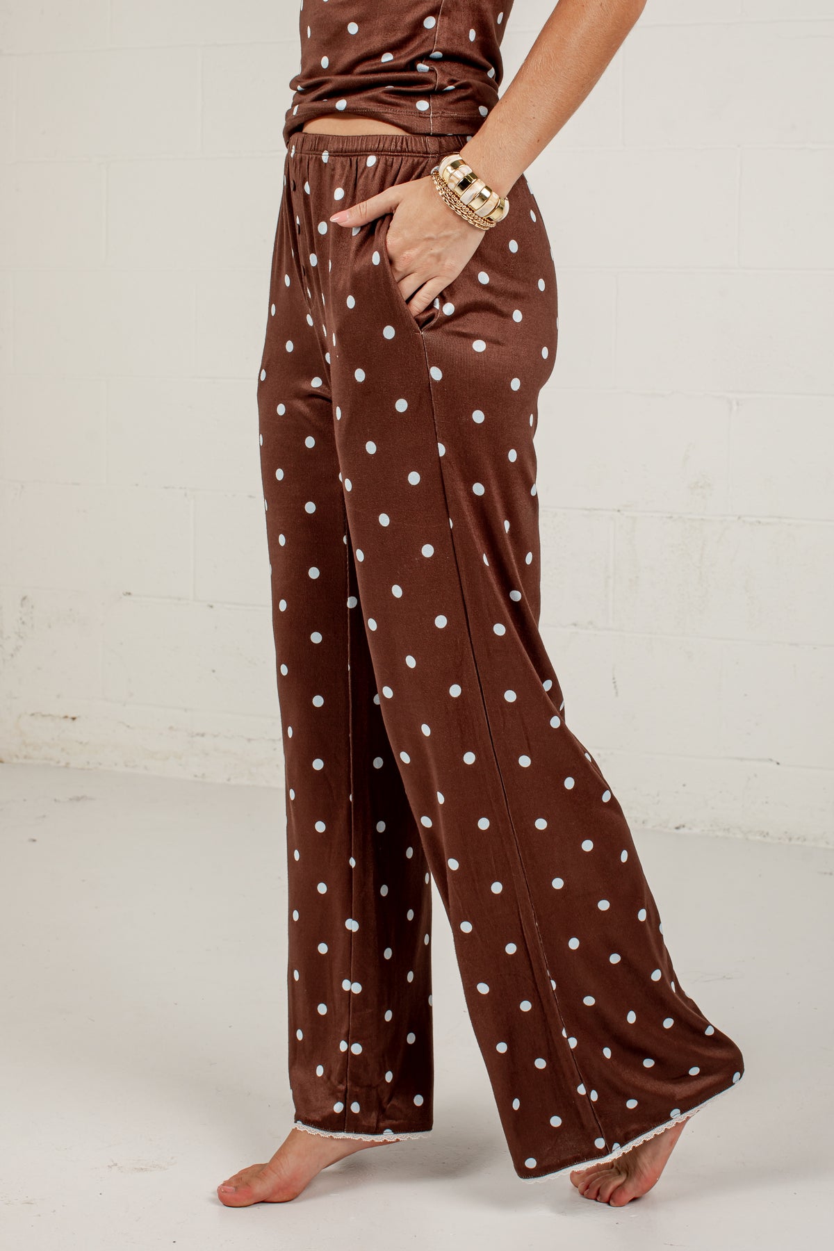 A person wearing brown wide-leg pajama pants with white polka dots, standing barefoot against a white wall.