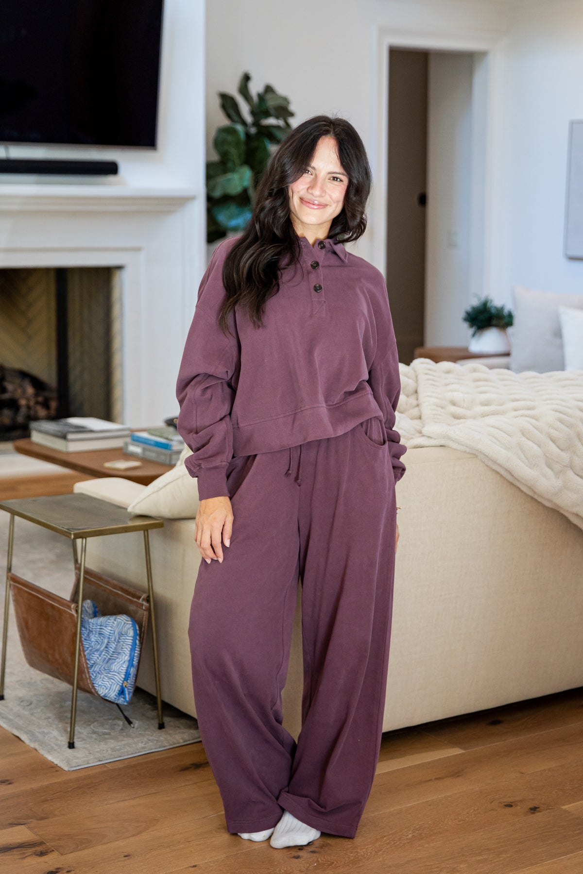 A woman wearing a matching mauve-purple lounge set consisting of a long-sleeve button-front top and wide-leg pants, standing in a cozy living room with wood floors, a bed, and a fireplace.