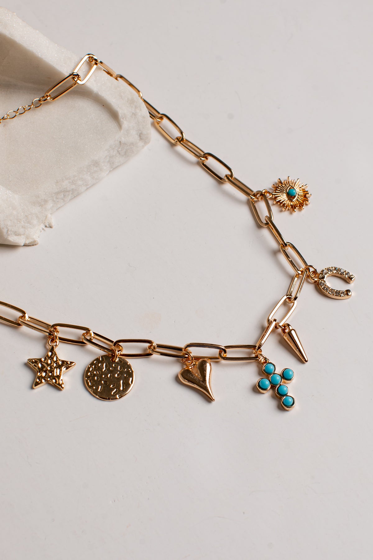 A gold-tone chain necklace with multiple charms, including a starfish, a round medallion, a crescent moon, a flower with turquoise accents, and a small sunburst charm, displayed on a light neutral surface.