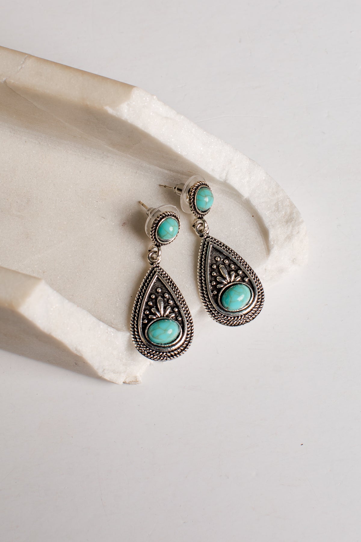 A pair of silver-toned teardrop dangle earrings with oval turquoise stones, displayed on a textured white stone prop against a light neutral background.