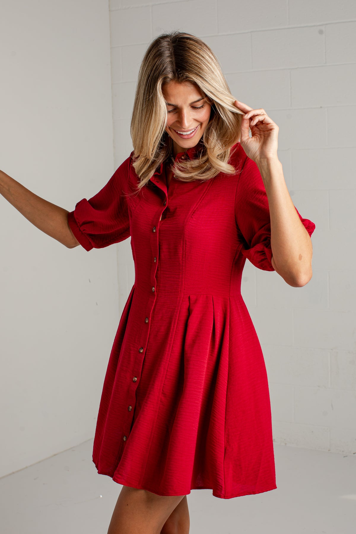 A woman modeling a bright red short-sleeve button-front dress with a flared skirt, standing against a light gray wall.