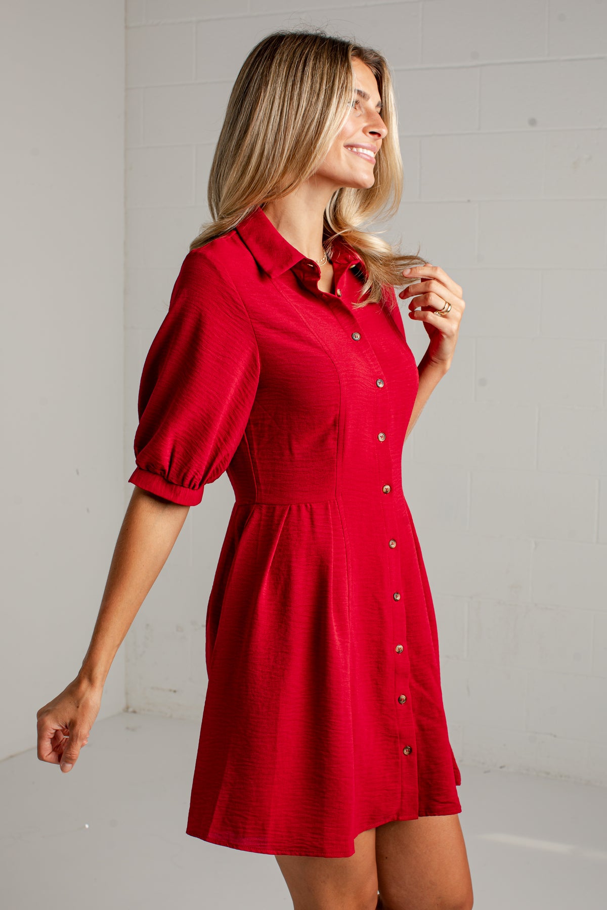 A woman wearing a bright red short-sleeve button-up dress with puff sleeves and a gathered waist, standing against a light gray wall.