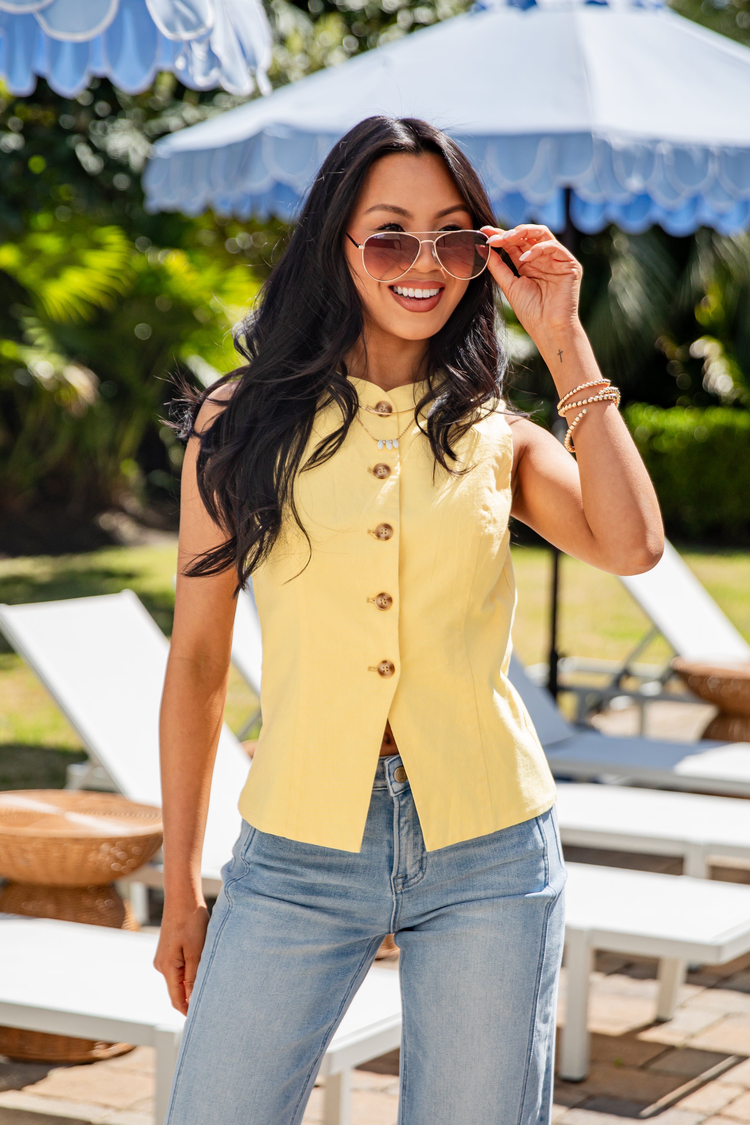 Woman wearing a yellow sleeveless top and blue jeans, standing outdoors with lounge chairs and umbrellas in the background.