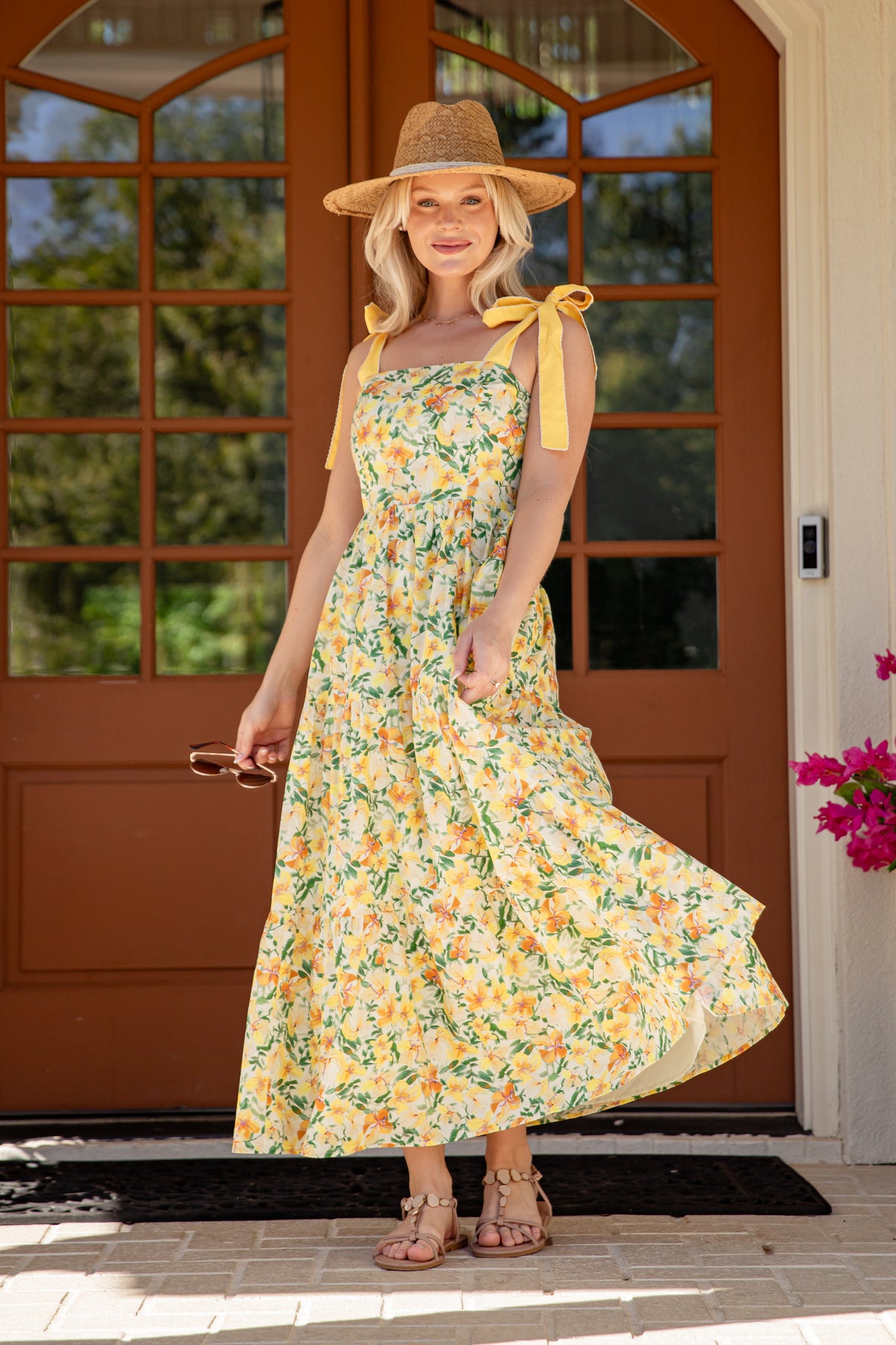 Woman in a floral dress and hat standing in front of a brown door with greenery.