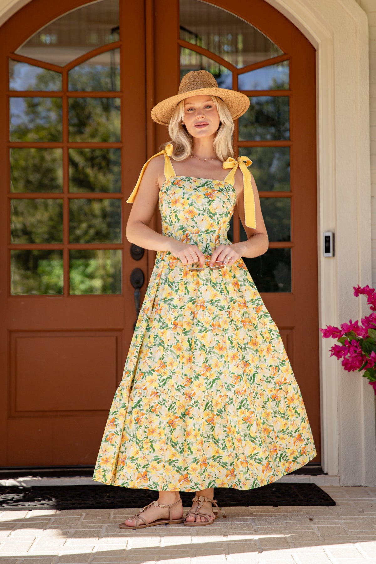 Woman in a floral dress and sun hat standing in front of a brown door with glass panels.