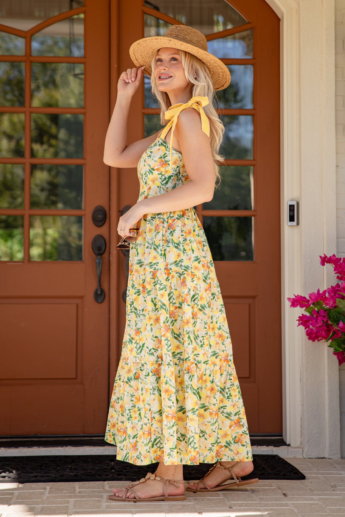 Woman in a floral dress and sun hat standing in front of a brown door.