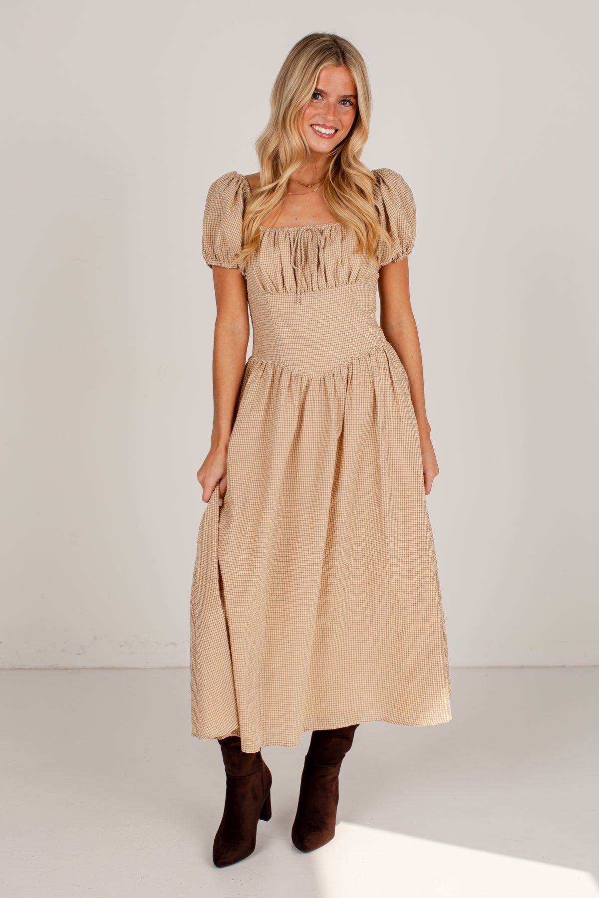 A woman modeling a beige midi dress with puff sleeves, a square neckline, and a gathered skirt, paired with dark brown ankle boots, against a plain white studio background.