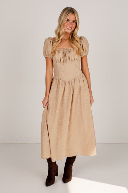 A woman modeling a beige midi dress with puff sleeves, a square neckline, and a gathered skirt, paired with dark brown ankle boots, against a plain white studio background.