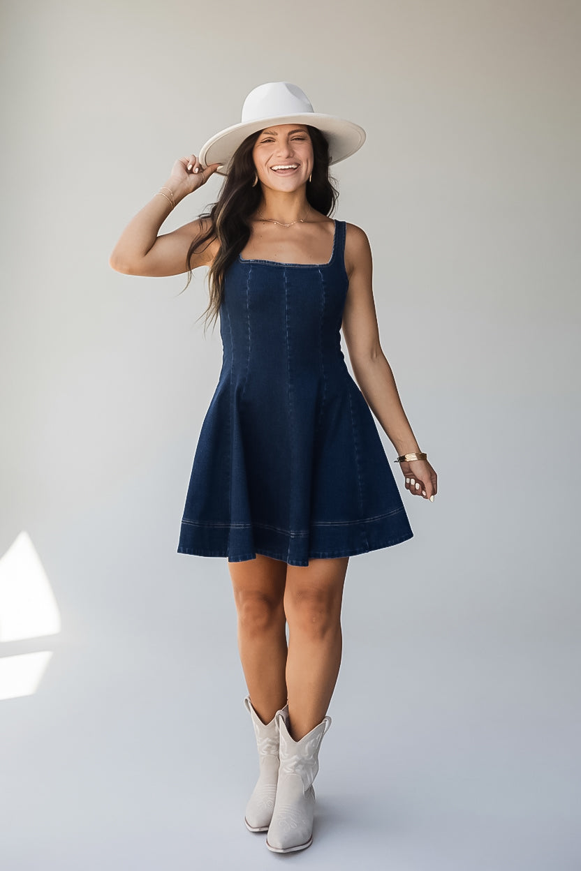A woman modeling a dark blue denim mini dress with a flared skirt, styled with a white wide-brim hat and white cowboy boots against a light gray studio background.
