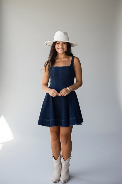 A woman modeling a dark blue denim dress with a white wide-brim hat and white cowboy boots, standing against a light gray studio background.