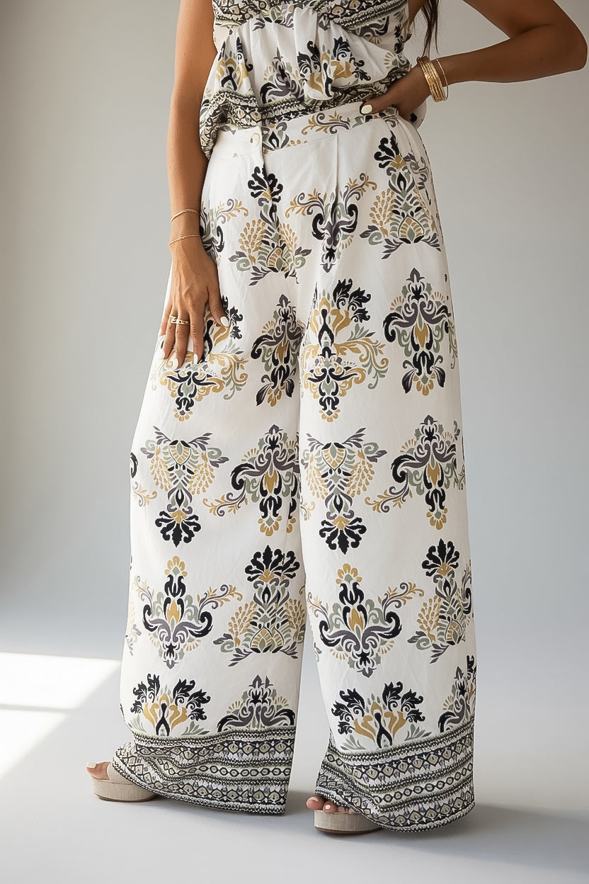 A model wearing a pair of wide-leg pants with a white base and an intricate black and gold damask-style floral pattern, paired with a matching patterned top.