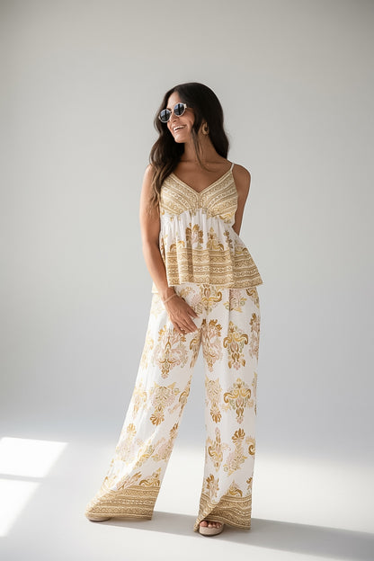 A woman modeling a matching sleeveless top and wide-leg pants set in a cream and gold paisley print, standing against a light gray studio background.