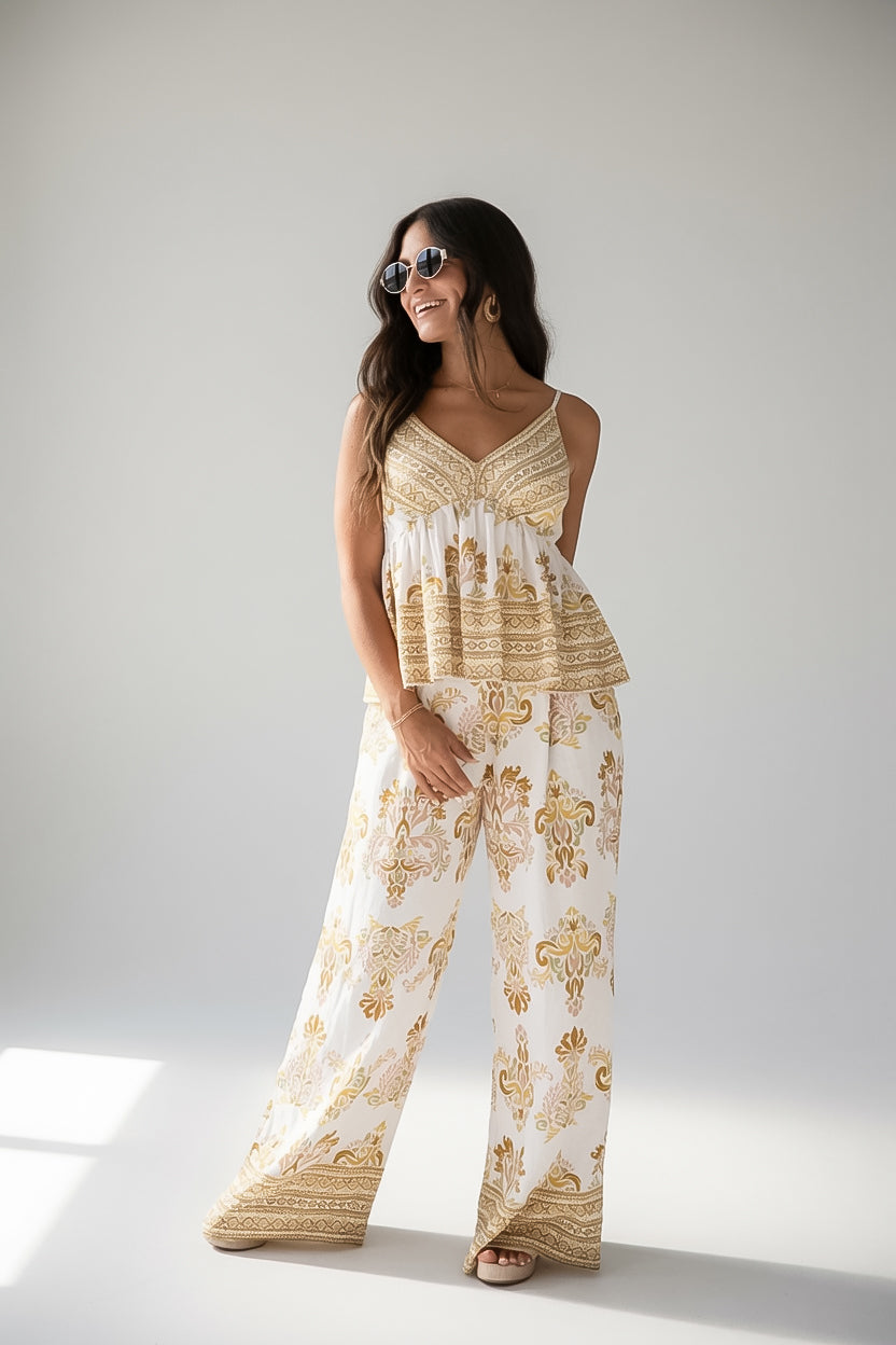 A woman modeling a matching sleeveless top and wide-leg pants set in a cream and gold paisley print, standing against a light gray studio background.
