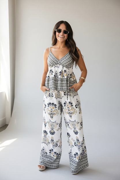 A woman modeling a sleeveless white jumpsuit with a navy and beige bohemian-style floral and geometric pattern, standing against a light gray studio background.