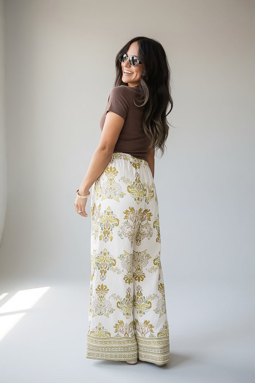 A woman wearing a brown short-sleeve top and high-waisted, wide-leg cream pants with a gold and green damask pattern, photographed in a studio setting.