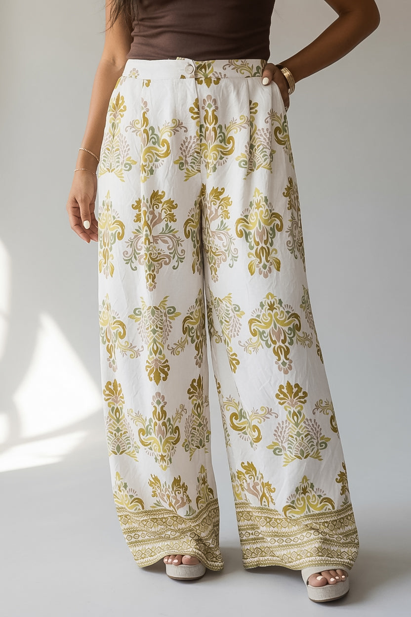 A person wearing high-waisted, wide-leg pants with an intricate white, gold, and olive green floral and paisley pattern, paired with a brown top and beige heels.