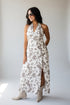 A woman modeling a sleeveless halter-neck maxi dress with a brown and beige floral pattern on a white background, paired with beige ankle boots, set against a plain light gray studio backdrop.
