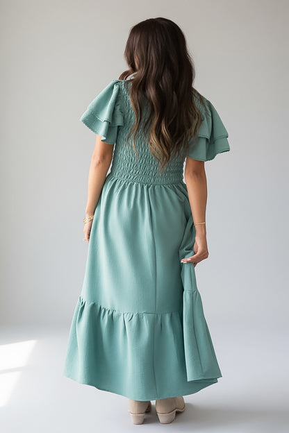 A woman viewed from behind wearing a sage green smocked maxi dress with ruffled short sleeves and a tiered hem, paired with beige heels, against a plain light gray studio background.