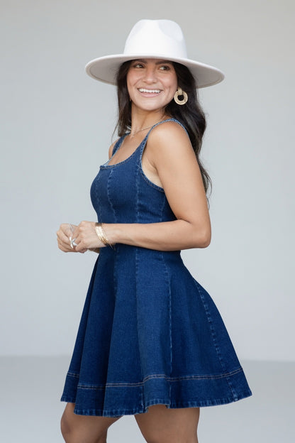 A woman modeling a dark blue denim mini dress with a flared skirt and a wide-brimmed white hat, posing against a light gray studio background.