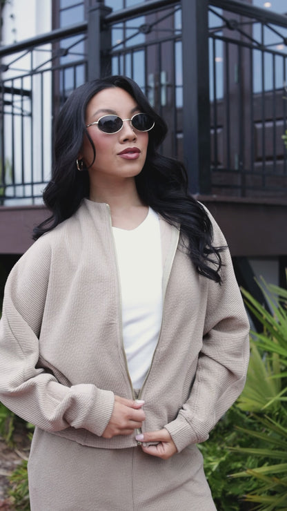Woman wearing a beige outfit standing outdoors with plants and a building in the background
