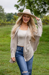 Woman wearing a beige hat, beige cardigan, white top, and blue jeans in a grassy outdoor setting.