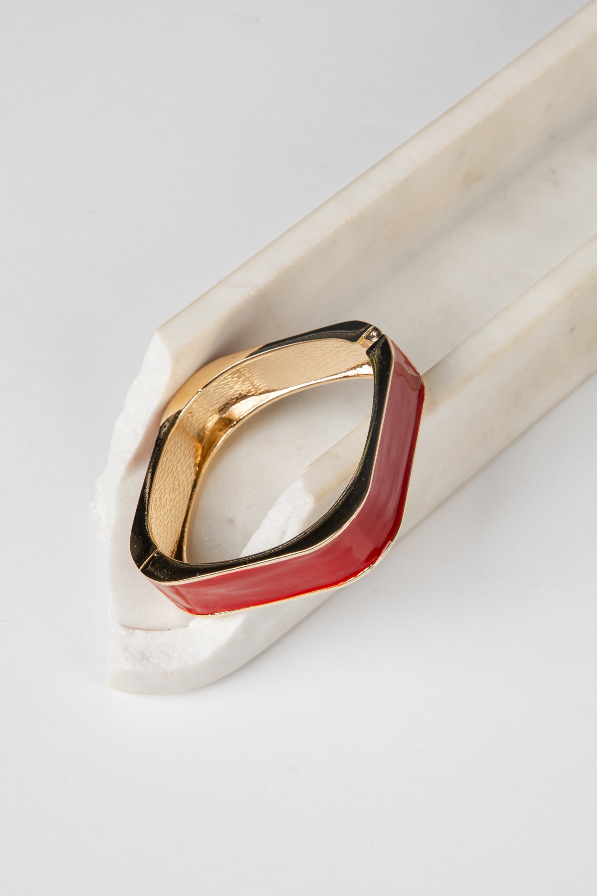 A geometric ring with a gold-toned metal band and a dual-tone red and black enamel inlay, displayed on a white marble stand against a light gray background.