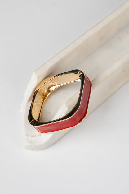 A geometric ring with a gold-toned metal band and a dual-tone red and black enamel inlay, displayed on a white marble stand against a light gray background.