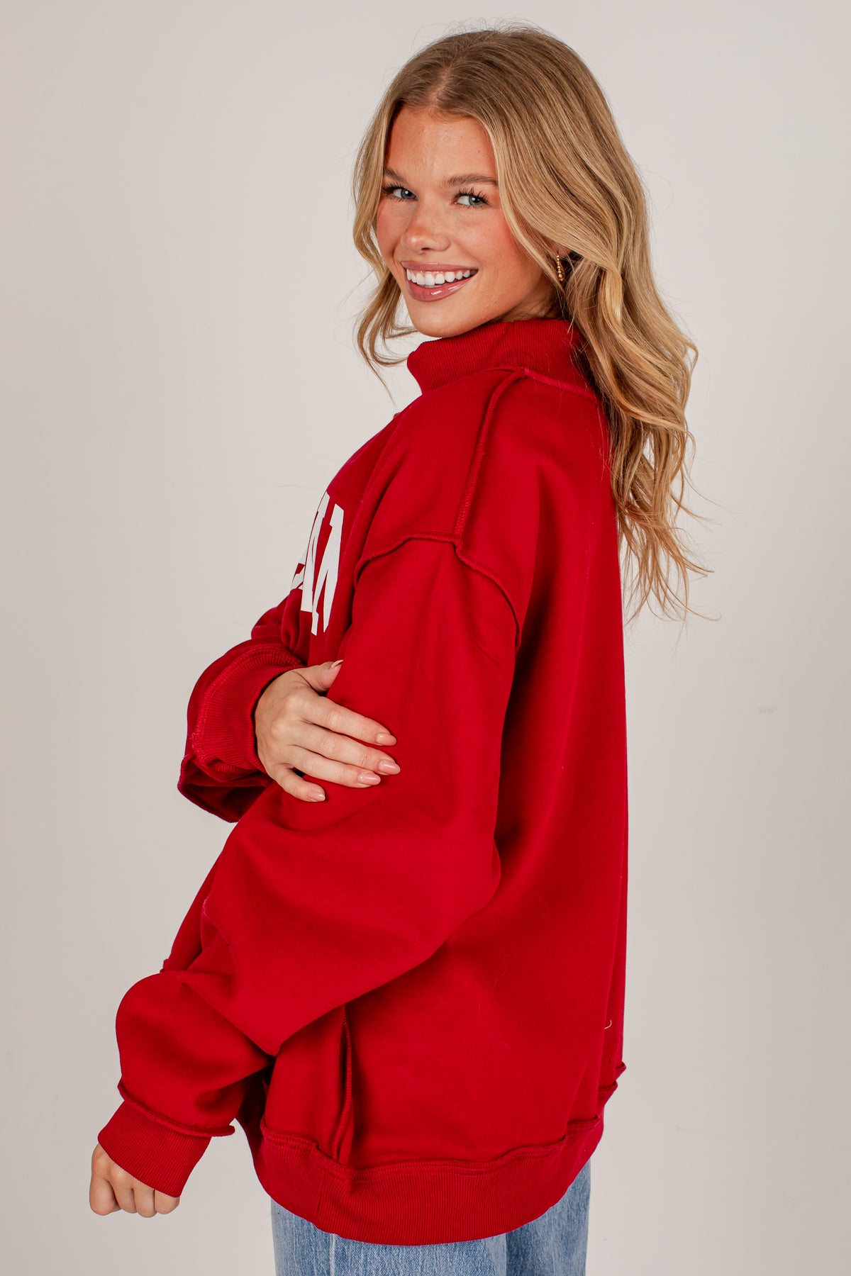 A woman with blonde hair wearing a red half-zip sweatshirt with white lettering on the back, paired with blue jeans, photographed against a light gray studio background.
